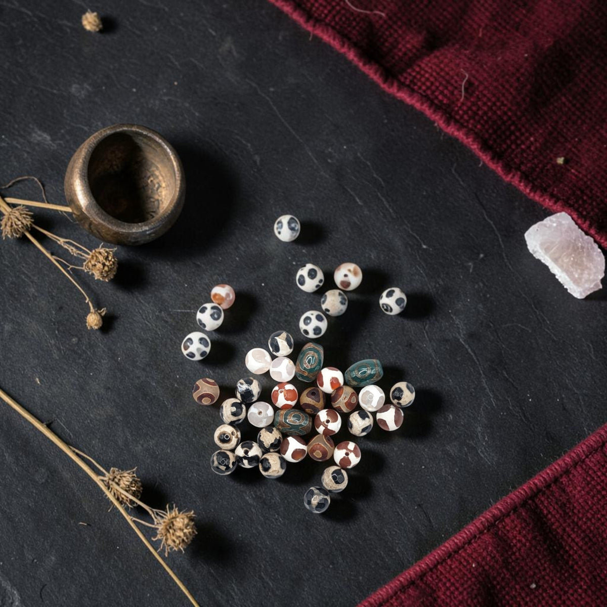 Tibetan Dzi-style agate beads with a bronze bowl, crystal, dried botanicals, and burgundy textile.